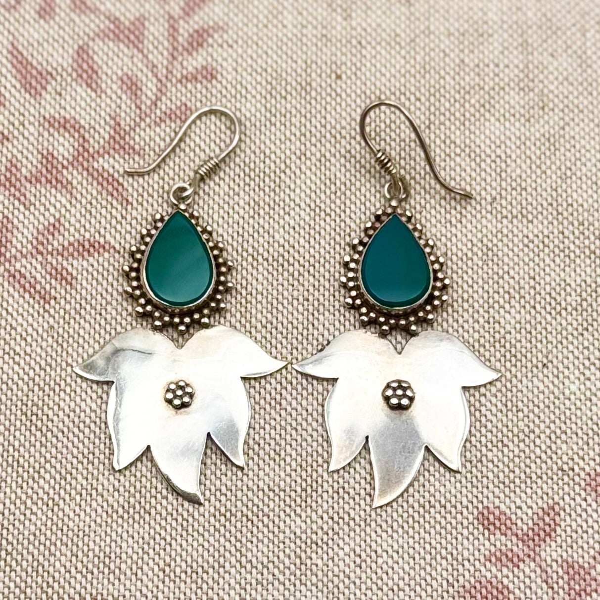 Pair of earrings with silver floral designs and teal teardrop stones on a textured fabric background.
