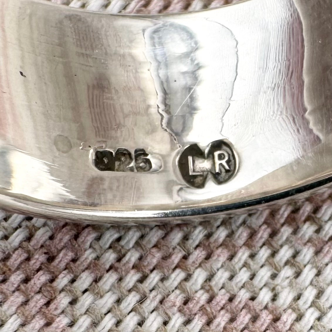Close-up of a silver ring with engraved design and visible hallmarks on a textured fabric background.