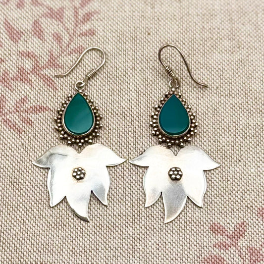 Pair of earrings with silver floral designs and teal teardrop stones on a textured fabric background.