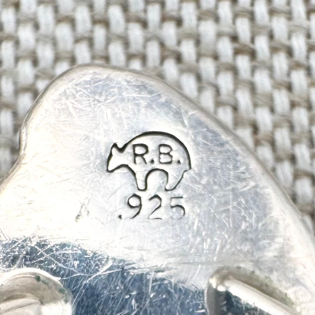 Close-up of a silver object with 'R.B.' and '925' stamp on a textured background