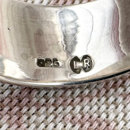 Close-up of a silver ring with engraved design and visible hallmarks on a textured fabric background.