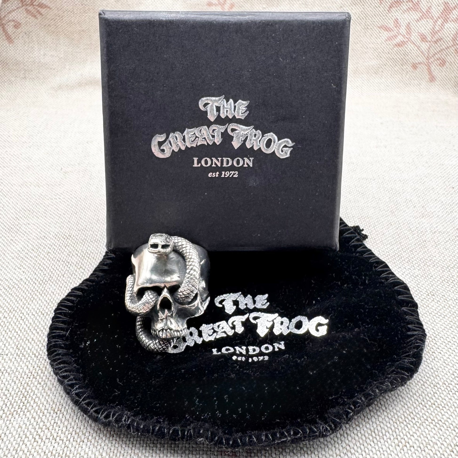 Silver skull and snake ring on a black cushion with 'The Great Frog London' branding.